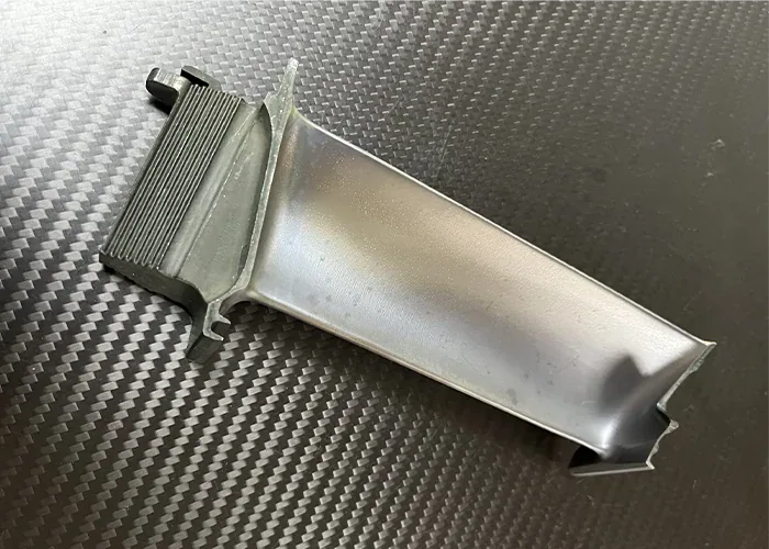Turbine blade blank sample on a carbon fiber background, showing the root dovetail and trailing edge